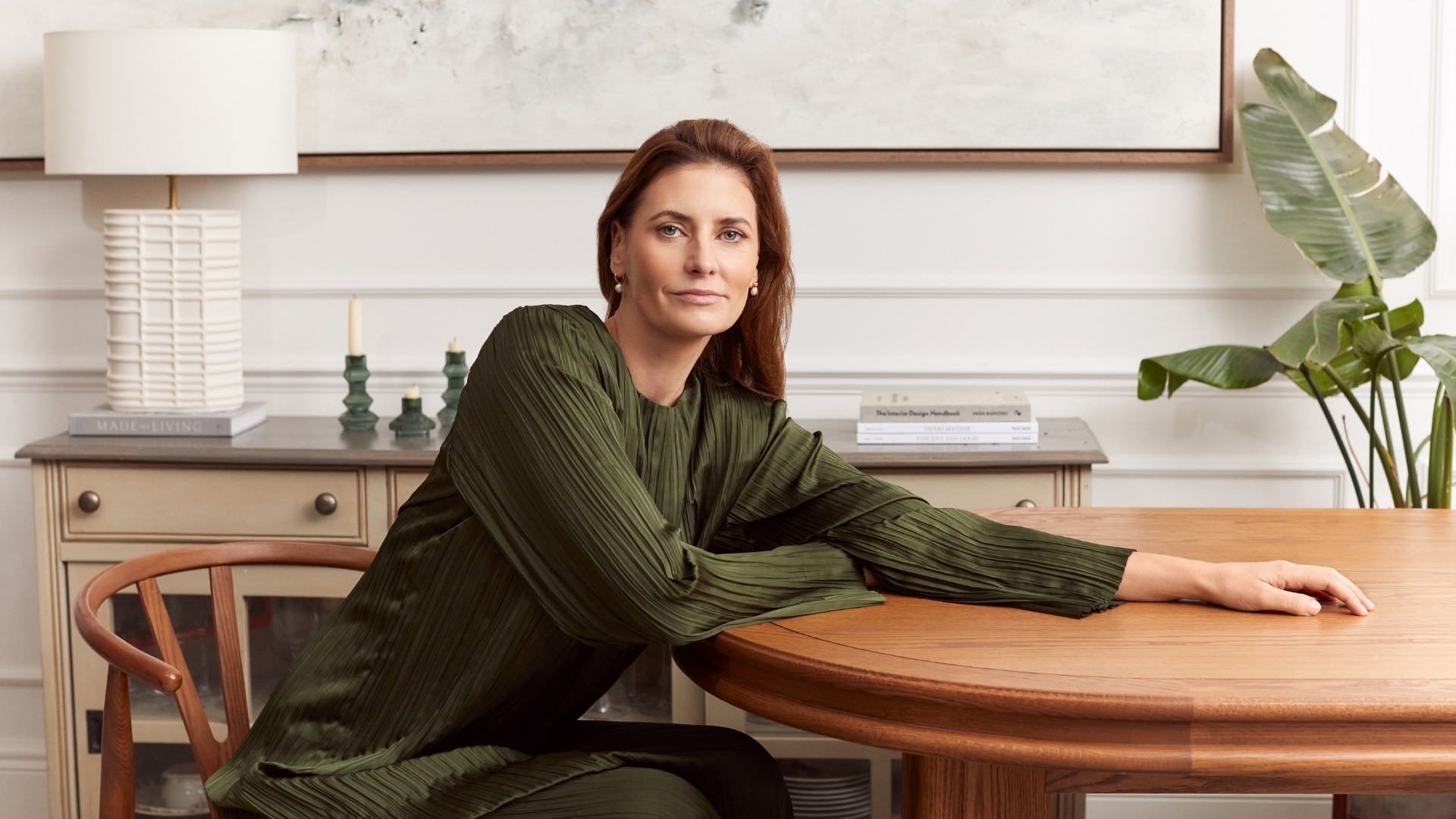 Woman with long auburn hair in a green jumper, seated at a wooden table at home