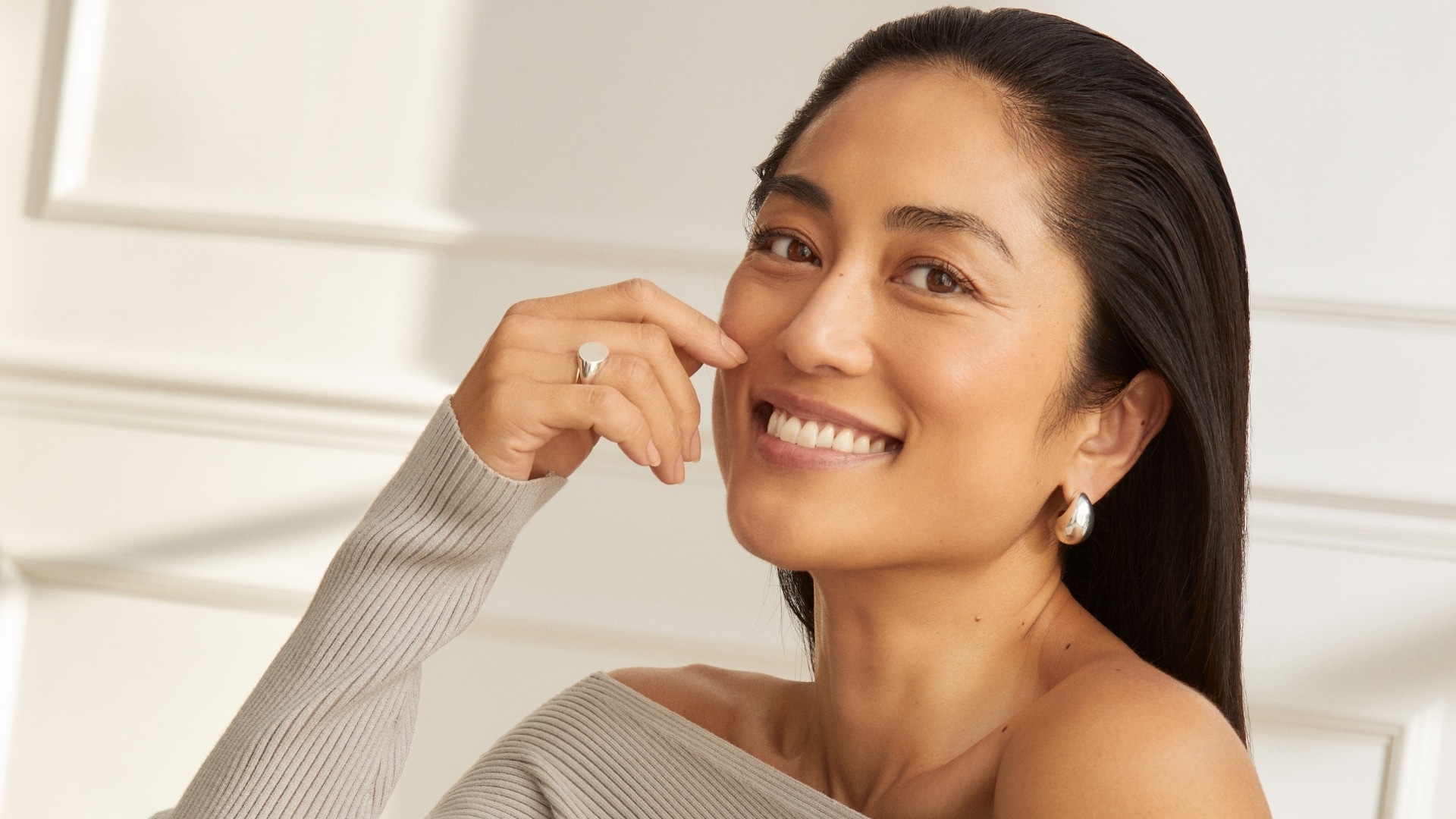 Smiling woman with dark hair touching her cheek, in a soft cream top