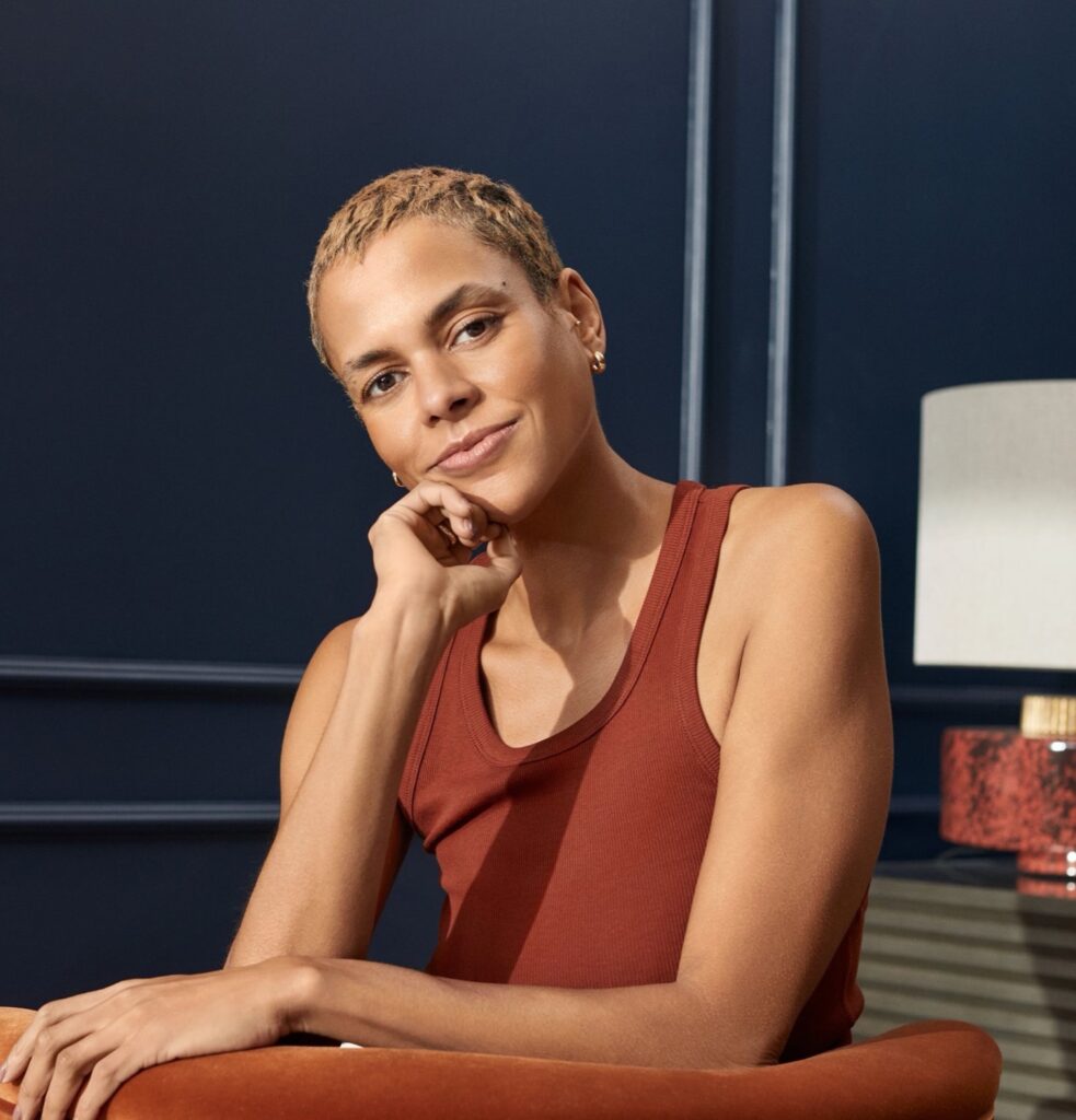 Woman with short blonde hair in a rust vest, resting her chin on her hand in a navy room
