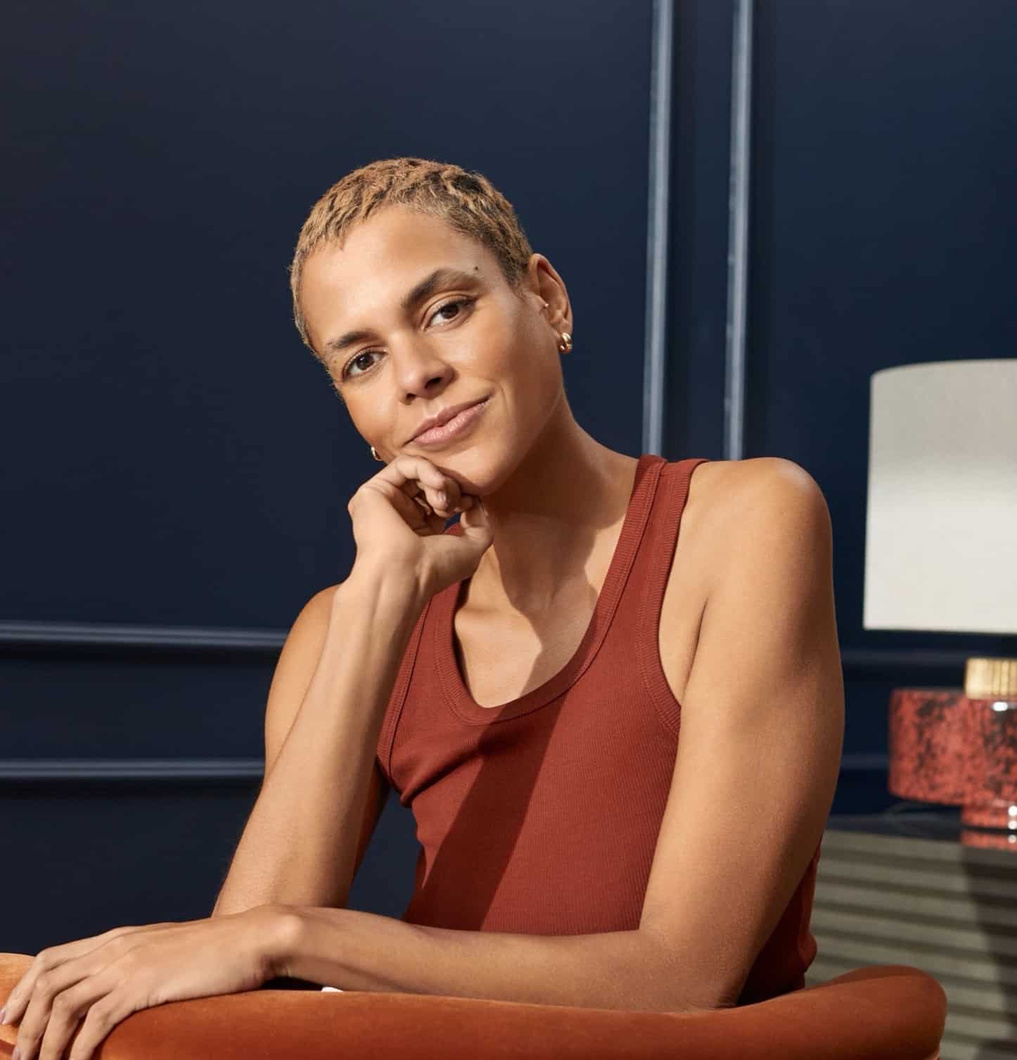 Woman with short blonde hair in a rust vest, resting her chin on her hand in a navy room