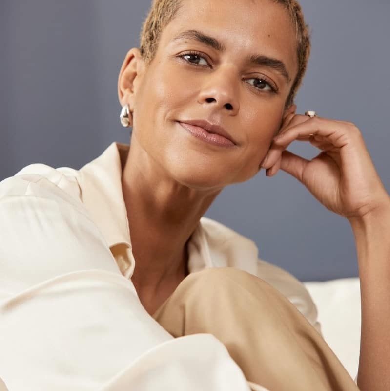 Woman with short cropped hair in a cream shirt, smiling with her hand resting against her cheek