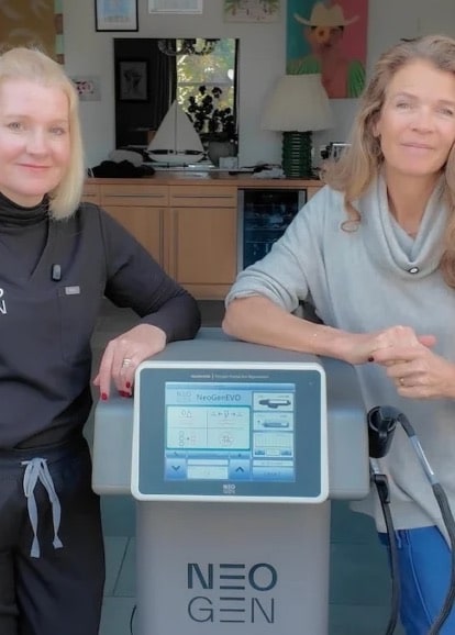 Two women standing either side of a NeoGen device with its touchscreen, in a bright living room