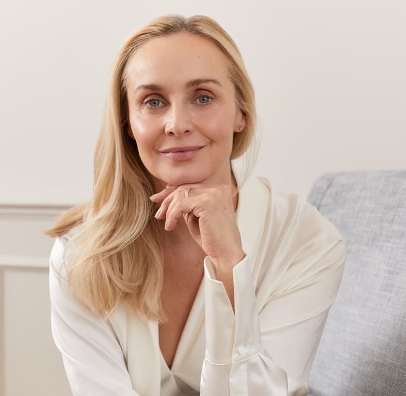 Woman with long blonde hair resting her chin on her hand, seated on a grey sofa in a cream room