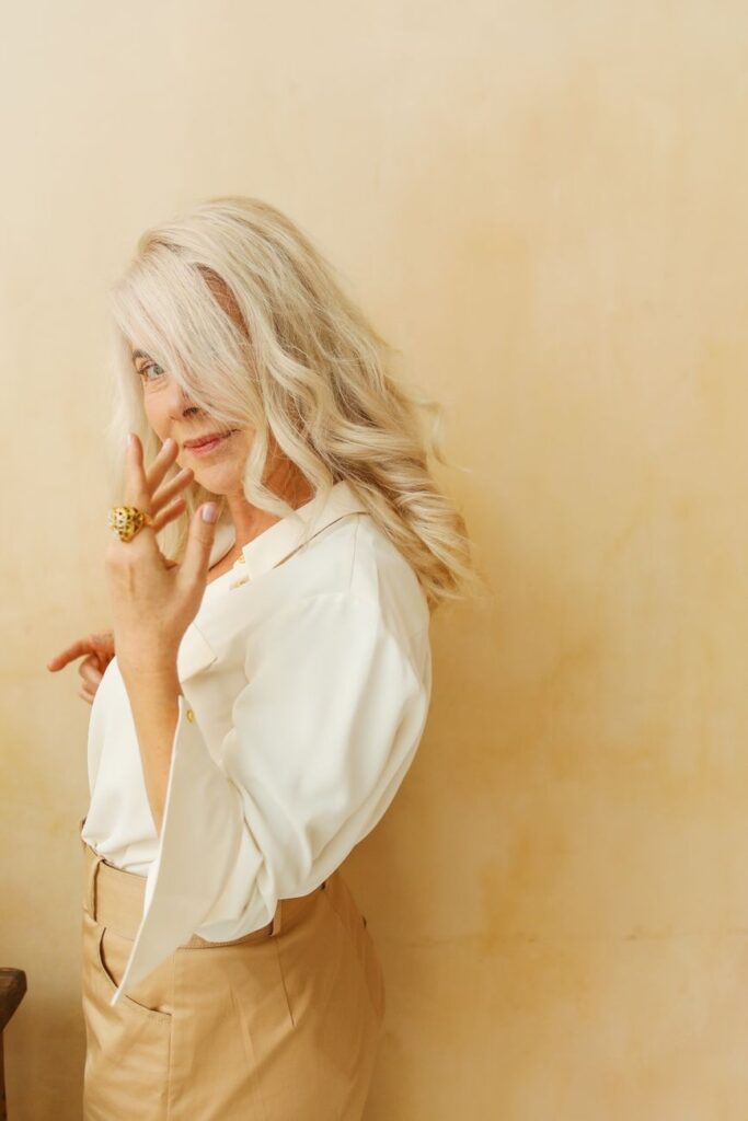 Woman with long blonde hair in a white shirt, smiling with a hand near her lips