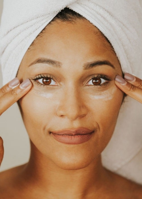 Woman in a white head towel smiling as she touches the skin beside her eyes