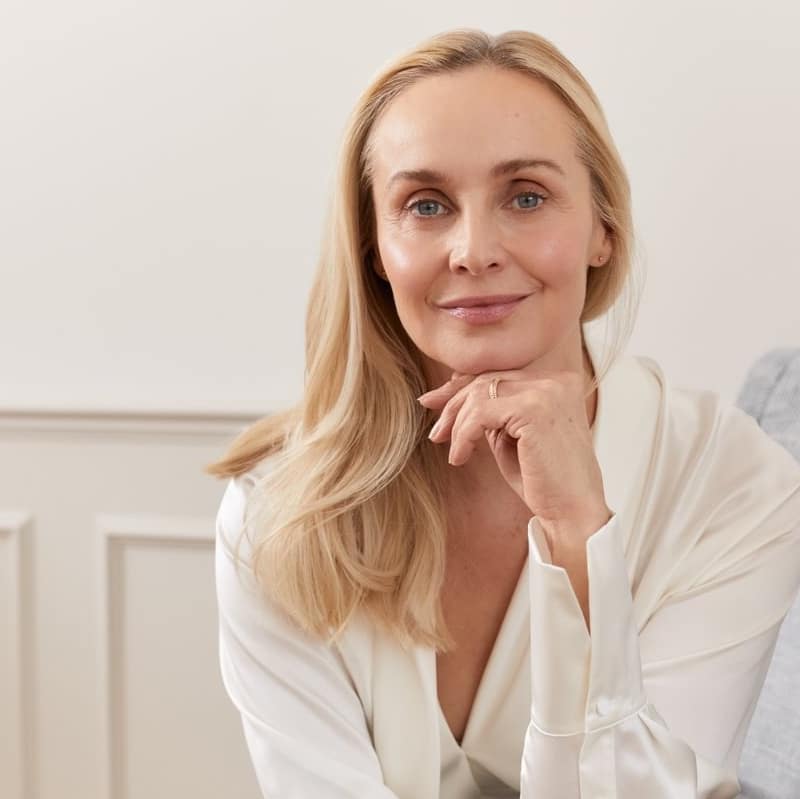 Woman with long blonde hair in a cream top, resting her chin on her hand on a grey sofa