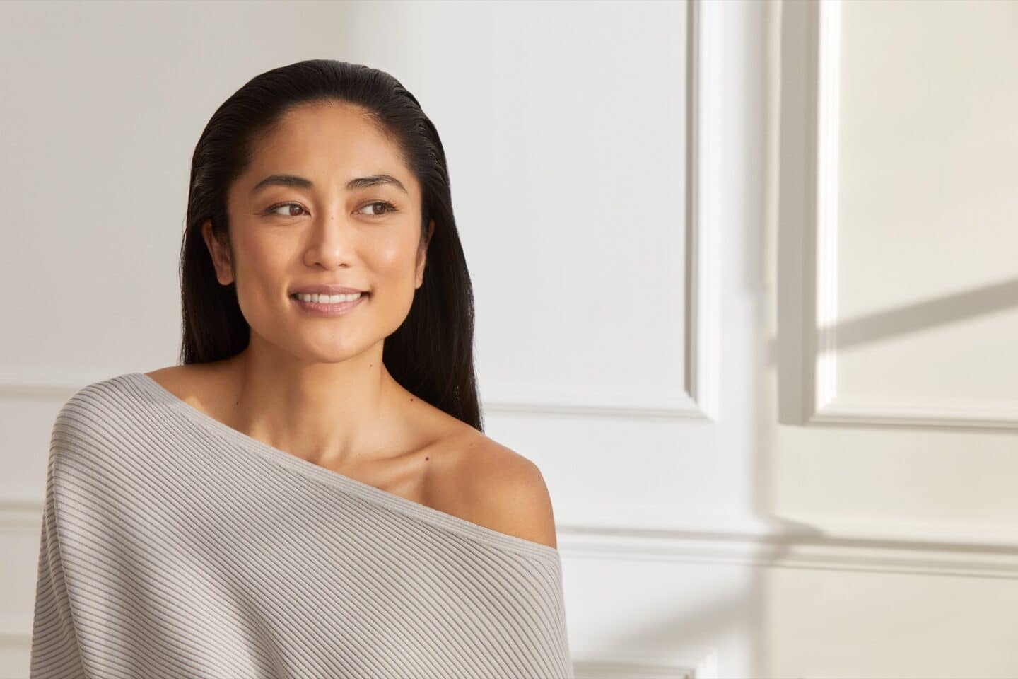 Smiling woman with long dark hair in a grey off-shoulder jumper, looking to one side
