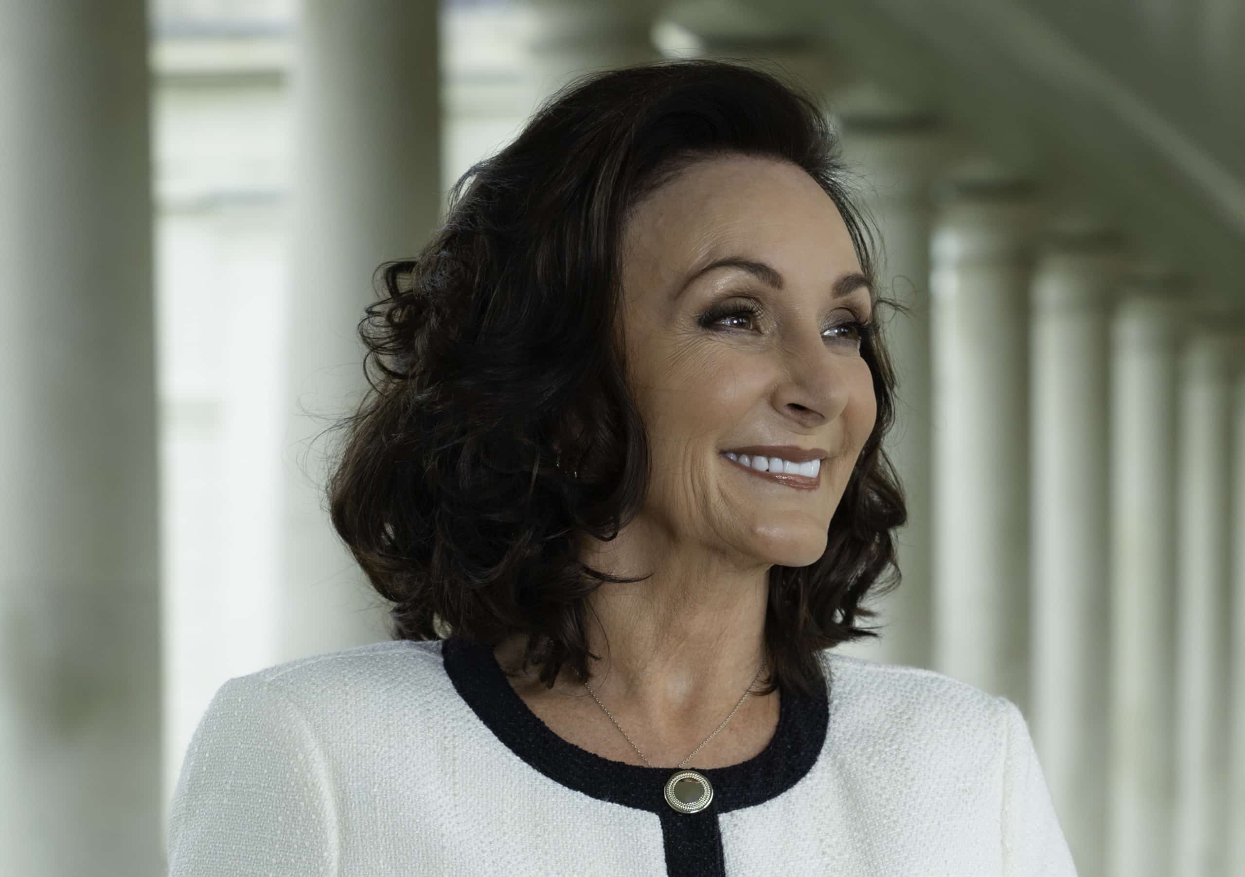 Shirley Ballas smiling in a white jacket, with a row of columns behind her